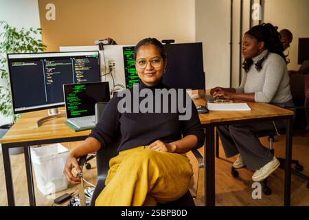 Portrait of confident businesswoman sitting with legs crossed at knee near desktop PC in tech startup office Stock Photo
