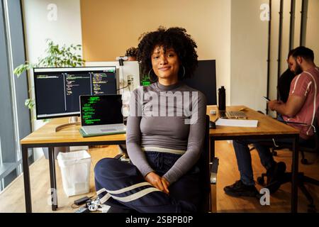 Portrait of curly haired female IT professional sitting in front of desktop PC at tech office Stock Photo
