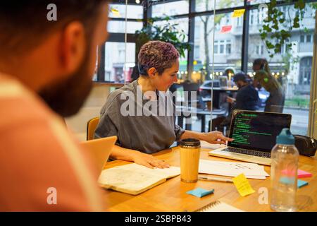 Non-binary IT professional discussing coding on laptop to colleagues during meeting in office Stock Photo