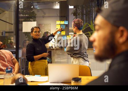 Non-binary business professional standing with female coworker pointing at adhesive note stuck on glass wall during meet Stock Photo