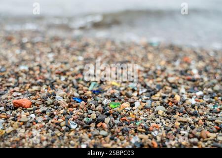 Close-up of various glass fragments on beach, mixed with sand and small stones, with gentle sea waves rolling in background Stock Photo