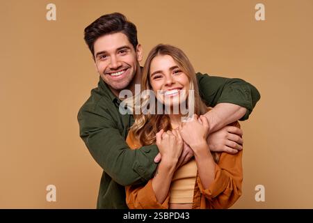 A happy and cheerful young couple are happily embracing each other. They radiate sincere smiles and are isolated on a warm beige background. Stock Photo