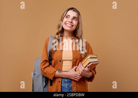A cheerful student with long hair beams joyfully while holding a stack of books. Dressed casually yet stylishly, she carries a trendy backpack, ready Stock Photo