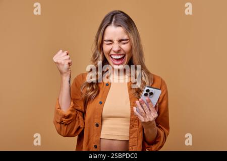 A cheerful young woman is happily holding her smartphone while wearing a cozy and trendy outfit. She stands against a soft brown backdrop that beautif Stock Photo
