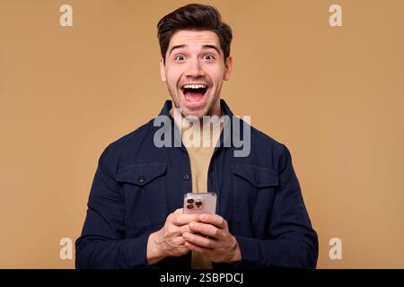 A joyful young man wearing a surprised expression has his eyes glued to his smartphone, his face radiating excitement. This image is perfect for theme Stock Photo