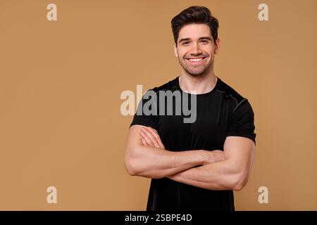 A cheerful young man with an engaging and confident smile stands proudly with his arms crossed against a warm beige backdrop, showcasing his relaxed a Stock Photo