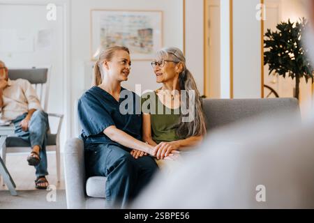 Female nurse talking with retired senior woman while sitting on sofa at nursing home Stock Photo