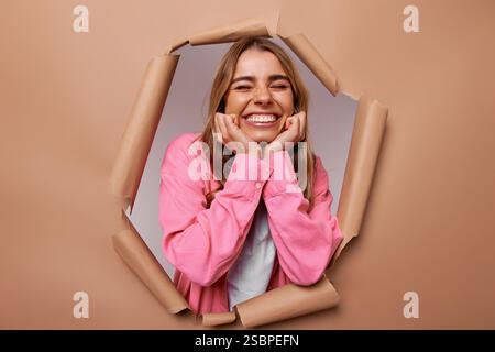 A vibrant young woman smiles widely and poses confidently, playfully peeking through a torn paper backdrop, embodying joy, creativity, and the delight Stock Photo