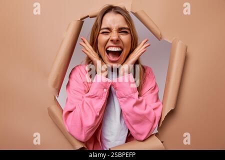 A vibrant young woman smiles widely and poses confidently, playfully peeking through a torn paper backdrop, embodying joy, creativity, and the delight Stock Photo