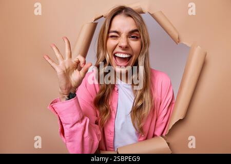A young woman joyfully appears from a torn brown paper backdrop, winking playfully and making an okay hand gesture, radiating vibrant energy and excit Stock Photo