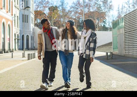 Three cheerful multi ethnic students are walking and talking together on a city street during a sunny winter day Stock Photo