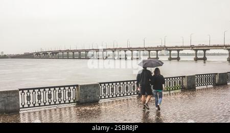 A couple walking along the river embankment under the umbrella in a rainy day. View on a long bridge through the river Stock Photo