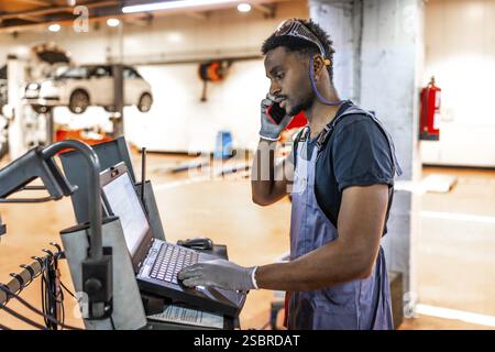Young mechanic using a laptop and talking on the phone while working on a car in a professional workshop Stock Photo