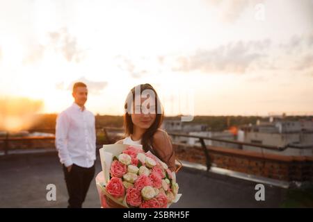 A young woman stands on a rooftop holding a bouquet of pink and white roses. Her partner watches her from a distance as the sun sets, creating a warm Stock Photo
