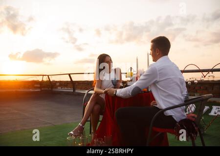 A couple sits at a beautifully set table on a rooftop during sunset, sharing drinks and engaging in conversation while the evening sky glows with warm Stock Photo