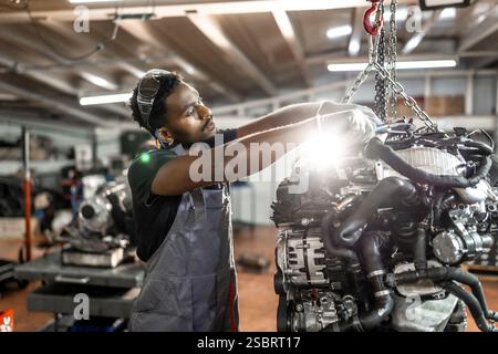 Skilled mechanic working diligently on a car engine while utilizing a crane in a modern auto repair shop, showcasing expertise and precision Stock Photo
