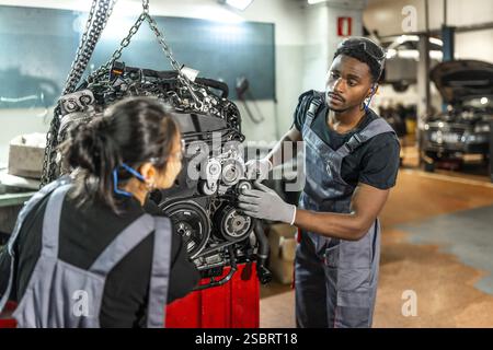 Two focused mechanics examining a car engine hanging from a crane in a professional workshop, collaborating on vehicle maintenance Stock Photo