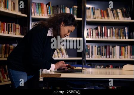 A dedicated woman stands studying on a laptop in a library, surrounded by shelves filled with books. The scene highlights focus, knowledge, and the pu Stock Photo