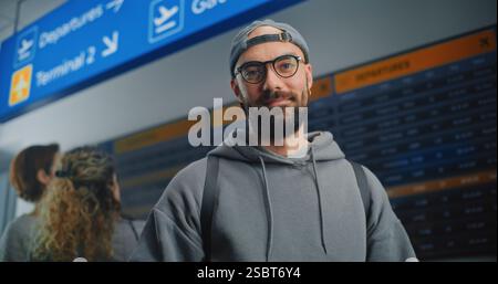 Crowded Airport Terminal: Man Scanning Plane Ticket Using Smartphone, Smiling, Looking at Camera. Digital Arrival and Departure Information Display in the Background. Traveler Going on Holiday Trip. Stock Photo