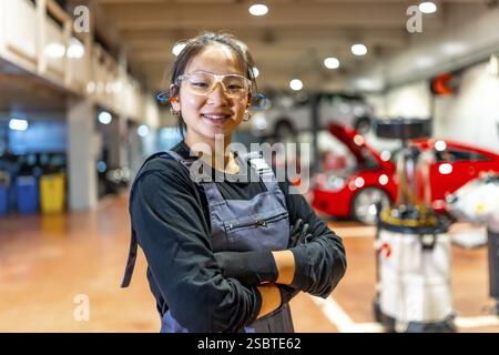 Confident young female mechanic wearing safety glasses and earplugs, smiling with crossed arms in a busy car repair shop Stock Photo