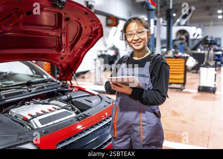 Confident female mechanic using a digital tablet, inspecting a car engine in a professional auto repair shop Stock Photo