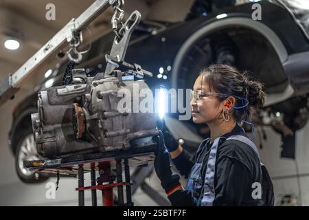 Young woman working as mechanic inspecting gearbox illuminated with flashlight in car workshop Stock Photo