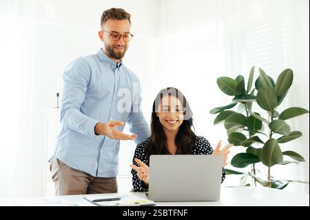 Colleagues working together. Professional young man in glasses leaning over a smiling brunette woman, pointing at a laptop screen during a business discussion in a modern office, teamwork concept Stock Photo