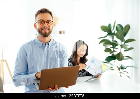 Positive successful arabian or caucasian businessman in a blue shirt and glasses, using a laptop browsing internet while standing in an office space, with a woman reviewing documents in the background Stock Photo