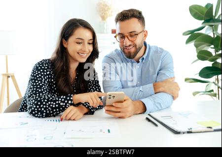 Cheerful multiracial colleagues sitting at a desk in office, looking at a smartphone together, discussing ideas, with charts and graphs scattered on the table and a modern laptop in front of them Stock Photo