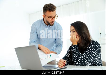Misunderstanding between coworkers. A worried man in glasses and a blue shirt shows financial chart to his female colleague, both looking at a graph with concerned expressions, analyzing reports Stock Photo