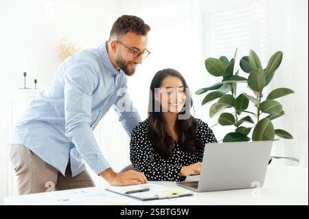 Two successful colleagues. Professional young man in glasses leaning over a smiling brunette woman, pointing at a laptop screen during a business discussion in a modern office, teamwork concept Stock Photo