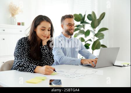 Concentrated young successful brunette business woman working at a desk in a bright modern office, takes notes on a documents, while her male colleague types on a laptop in the background Stock Photo