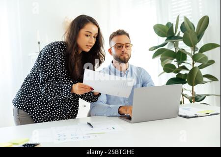 Concentrated business woman showing a business document to a male colleague in a modern office, both focused on a work with a laptop and charts on the desk, the sharing ideas, develop business plan Stock Photo