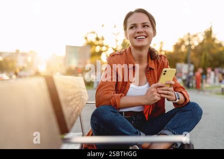 Young adult woman wearing a brown casual shirt sitting on a street bench and using her smart phone. Stock Photo