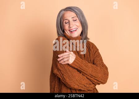 Stylish elderly woman embracing herself with a warm smile against a beige background, wearing a brown sweater Stock Photo