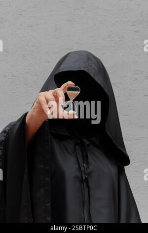 The unknown man in the black cloak points to the hourglass as a symbol of time Stock Photo