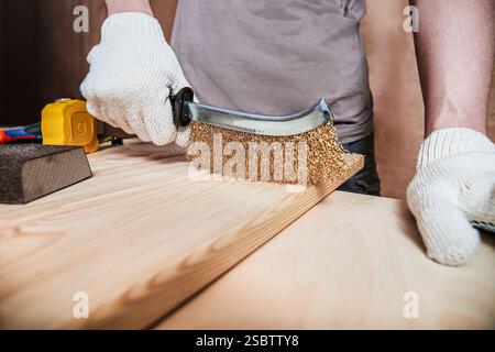 A person in gloves holds a brass wire brush, scrubbing a wooden board to create grain and rough wood texture Stock Photo