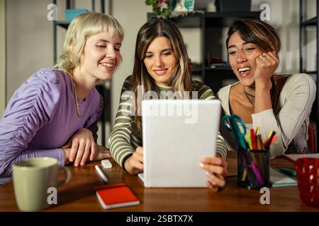 Three young women collaborating in a cozy, creative workspace. They are looking at a notebook, sharing ideas, and studying together, symbolizing teamw Stock Photo