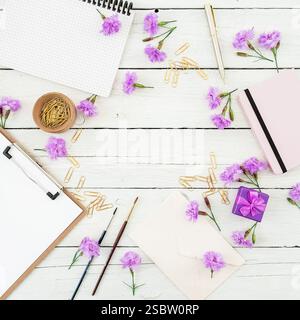 Flat lay of creative workspace with notebooks, brushes, purple flowers, gold paper clips, and gift box on white wooden background. Stock Photo