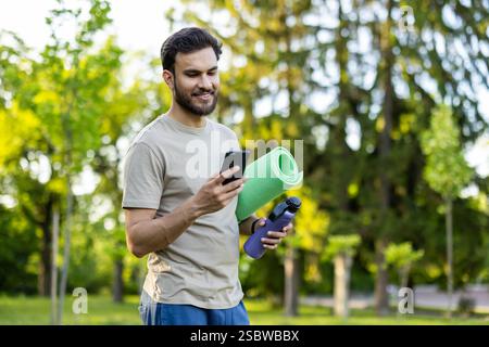 A young Indian man is jogging in the park, doing sports, holding a matchstick, a bottle of water and looking at the screen of a mobile phone. Stock Photo