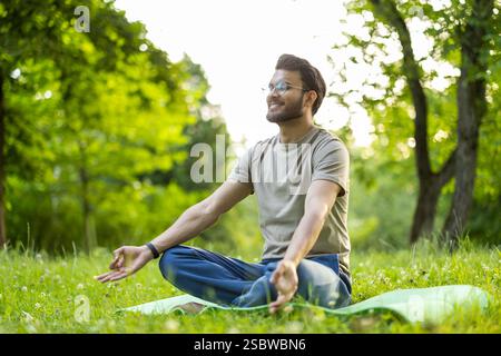 A smiling and calm young Indian man is sitting in the park on a mat in the lotus position, with his eyes closed and doing yoga.. Stock Photo