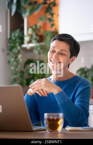 Happy smiling woman enjoy tea while make video call on laptop at home. Online learning, virtual meet Stock Photo