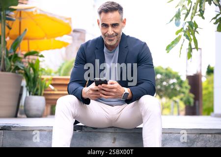Smiling Man Using Smartphone While Sitting Outdoors in Urban Setting Stock Photo