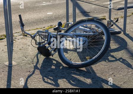 Abandoned and damaged bicycle lying on the ground near a metal rack on a city street in sunlight. Urban decay, vandalism and abandoned property Stock Photo