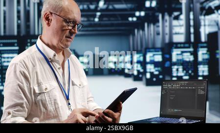 IT solutions expert in data center environment, solving tasks on tablet while running scripts on laptop terminal. Skilled specialist amidst servers, accessing databases and manipulating lines of code Stock Photo