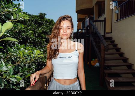 A cheerful young woman poses confidently outdoors by a wooden staircase, surrounded by lush greenery under the bright sun Stock Photo