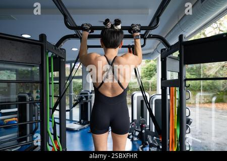 Strong female athlete exercising back muscles by doing pull ups on a horizontal bar in a well equipped fitness center, demonstrating strength and dedi Stock Photo