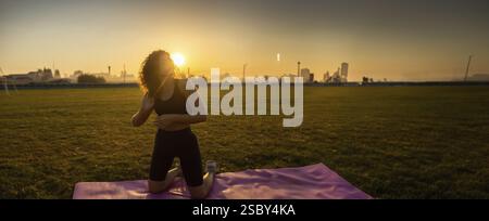 A woman kneels on a yoga mat, preparing for exercise, with the sunrise in the background Stock Photo