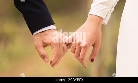 Hands of a newlywed couple, holding hands by little finger, with husband and wife rings, blurred background, white wedding dress Stock Photo
