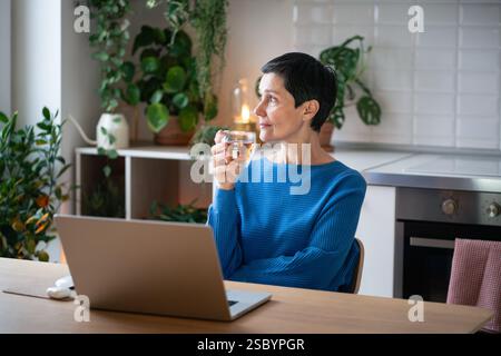Calm thoughtful mature woman enjoying cup of tea during break from working remotely on laptop Stock Photo